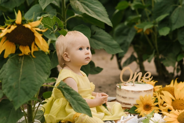 ひまわり畑でケーキと小さな美しい幸せな1歳の女の子の肖像画