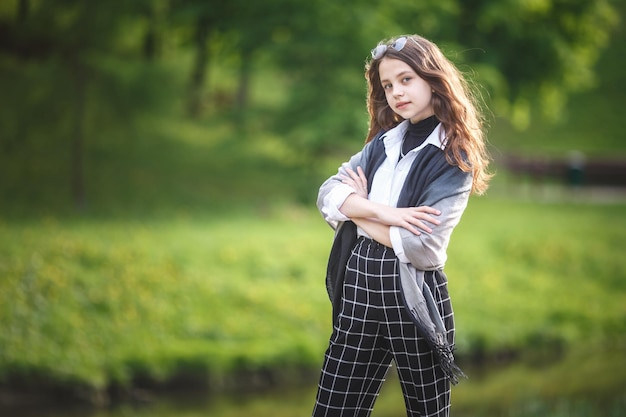 Portrait of little beautiful stylish kid girl with sunglasses and short plaid pants in city park on green forest background