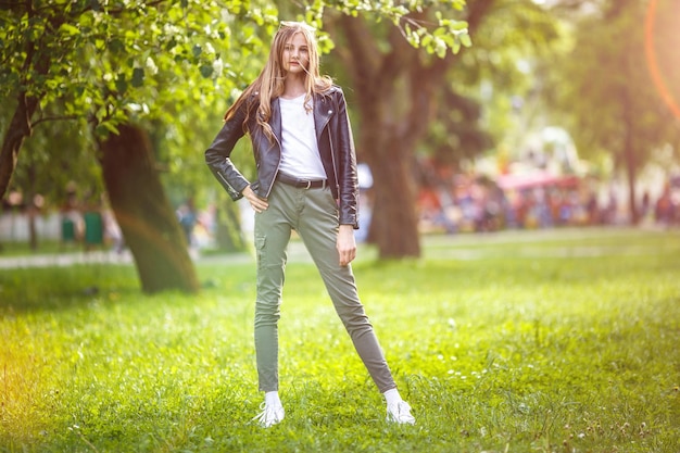 Portrait of little beautiful stylish tall kid girl with long hair in leather black jacket in city park on green forest background