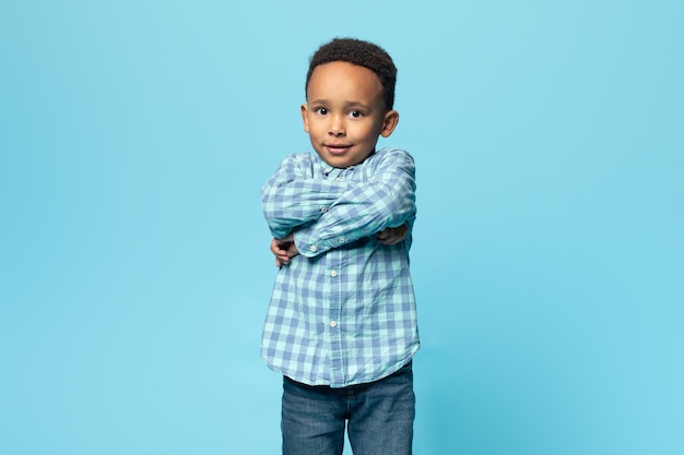 Portrait of little black boy standing with folded arms and looking at camera posing over blue