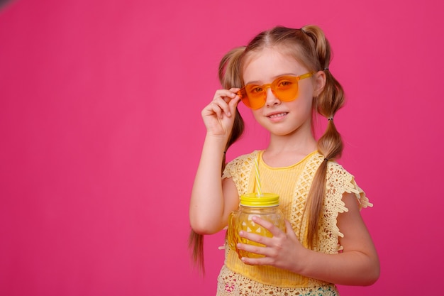 Portrait of a little blonde girl in sunglasses on pink with a glass jar of cooling cocktail.