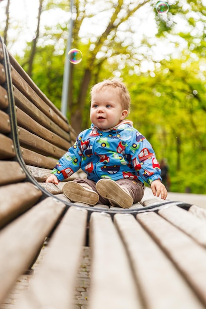 Портрет маленького мальчика в парке на скамейке, ловящего мыльные пузыри