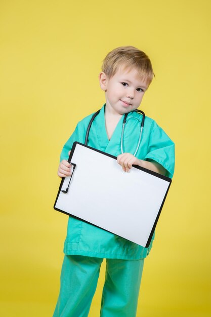 Portrait of little caucasian boy dressed in doctors green coat holds tablet in hand isolated on yellow