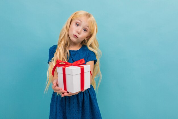 Il ritratto di piccola ragazza caucasica in vestito blu con capelli biondi lunghi tiene la scatola bianca con un regalo isolato su fondo blu