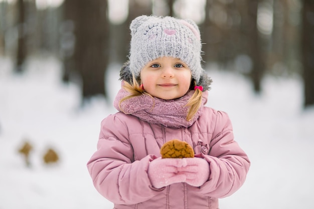 Ritratto di bambina, in posa all'aria aperta nella foresta innevata invernale con un gustoso biscotto fatto in casa nelle mani.