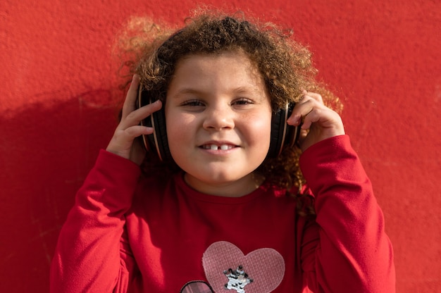 Portrait of little curly girl looking at camera wearing headphones. Isolated on red background.