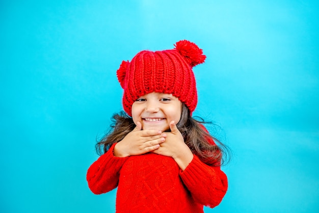 Portrait of a little curly girl in a red knitted hat in winter on a blue background Place for text