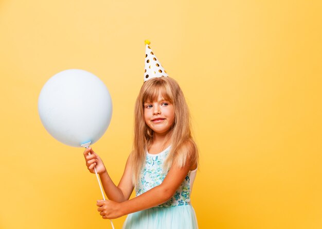 Portrait of a little cute girl in a festive cap and with balloons on a yellow background.
