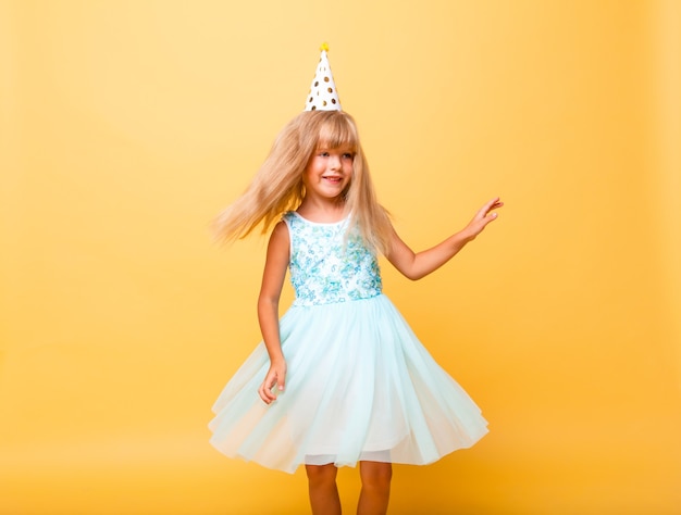 Portrait of a little cute girl in a festive cap and with balloons on a yellow background.
