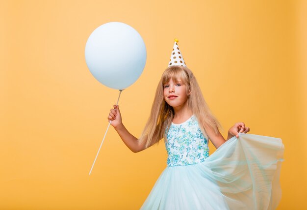 Portrait of a little cute girl in a festive cap and with balloons on a yellow background. 
