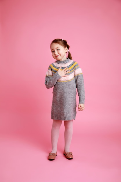 Portrait of little funny smiling girl showing to themself in warm dress on rosa background.