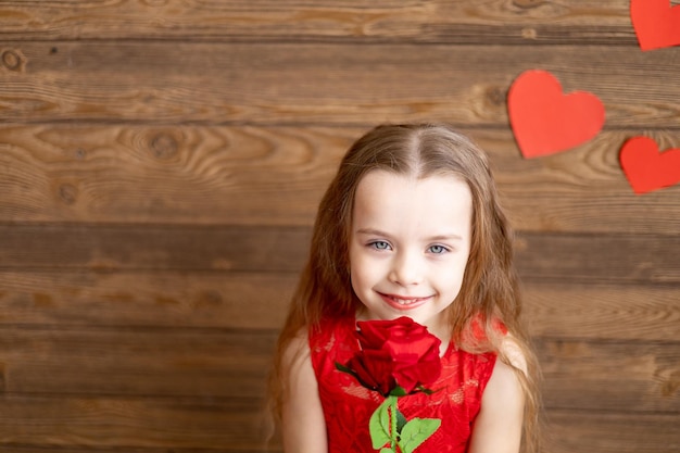 暗い茶色の木製の背景に赤いバラを保持し、バレンタインデーの概念をテキスト用の空きスペースに優しく微笑む赤いドレスを着た少女の子供の肖像画