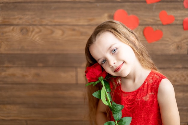 Portrait of a little girl child in a red dress holding a red rose on a dark brown wooden background and smiling sweetly the concept of Valentines day an empty space for text