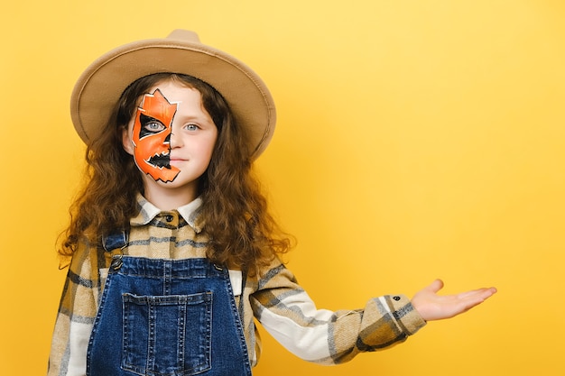 ハロウィーンの化粧マスクを持つ小さな女の子の肖像画は、スタジオで黄色の背景の上に孤立してポーズをとって、製品またはテキストのコピースペースで開いた手のひらを示しています。パーティーの休日のコンセプト
