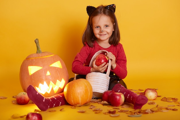 Portrait of little girl in costume of cat, kid sitting on floor with trick or treat basket, surrounded with apples and Jack o Lantern