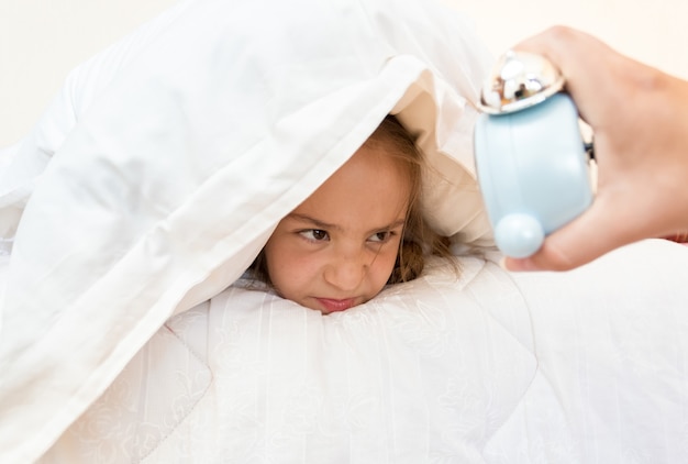 Ritratto di bambina che copre la testa con il cuscino e guarda la sveglia