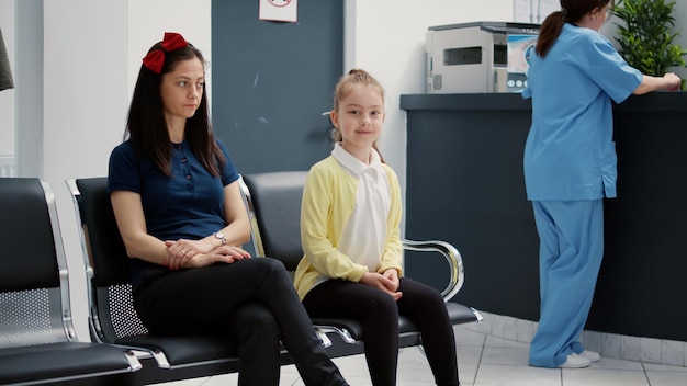 Portrait of little girl and mother sitting in waiting room area at medical clinic, waiting to attend consultation appointment with general practitioner. Hospital reception with diverse patients.