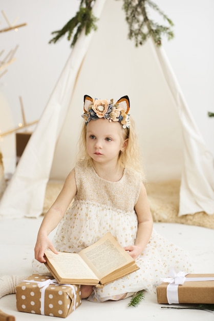 A portrait of a little girl near the wigwam and Christmas tree unpacking Christmas gifts on a white background