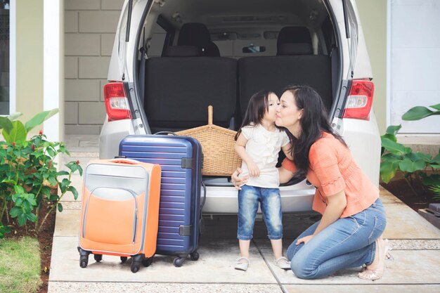 家のガレージで撮影された車の近くで母親にキスをしている中,休暇旅行の準備をしている小さな女の子の肖像画