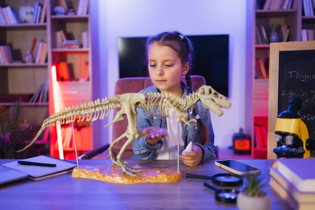 Portrait of little girl sitting at table and examine skeleton of dinosaur