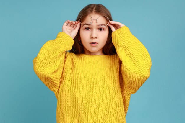 Portrait of little girl takes off glasses and looking at camera\
with astonishing facial expression, wearing yellow casual style\
sweater. indoor studio shot isolated on blue background.