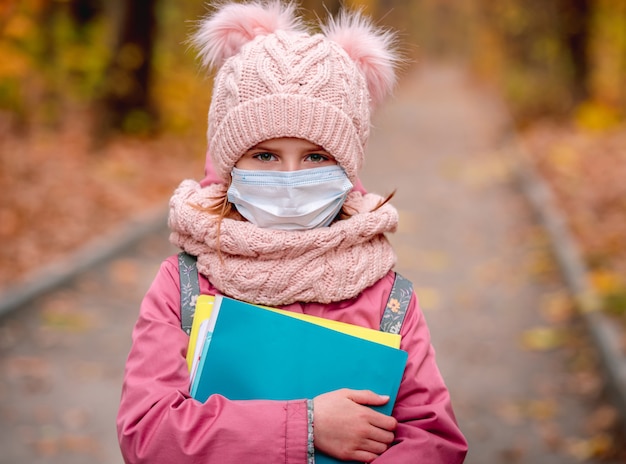 Portrait of little girl wearing protective face mask during walk through autumn park after school