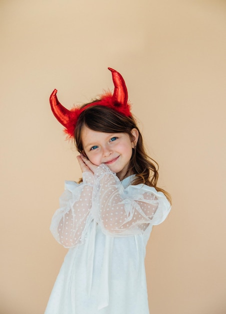 Portrait of a little girl in a white dress with devil horns Halloween costume Angelic facial expression The concept of two different personalities in character