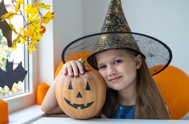 Portrait of little girl in witch costume holding pumpkin with Jack O Lantern face. Kid in black hat