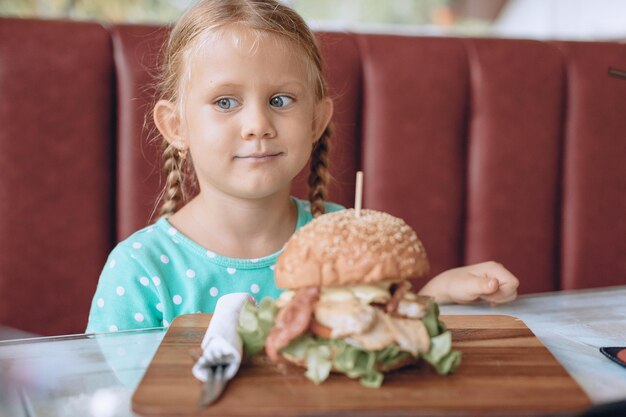 Photo portrait of a little girl with blond hair and blue eyes sitting next to the wooden tray with a huge tasty burger. fast food concept.