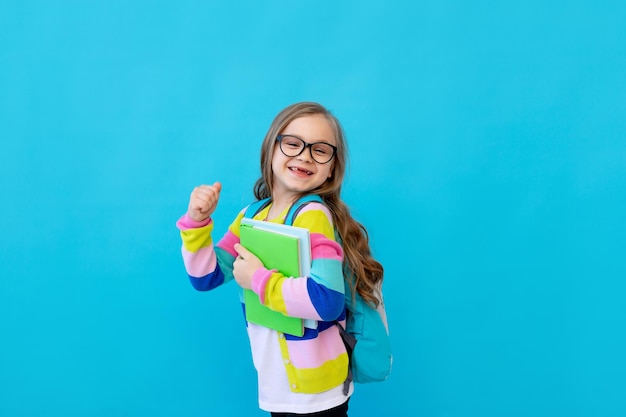Портрет маленькой девочки в очках в полосатой куртке с тетрадями и учебниками в руках и рюкзаком Концепция образования Фотостудия синий фон место для текста