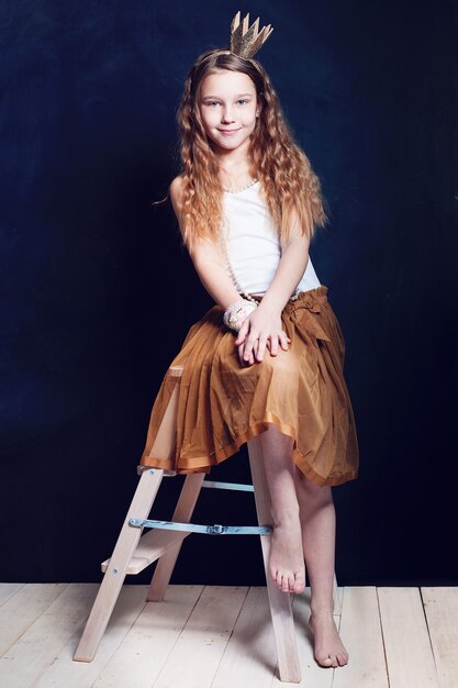 Portrait of little girl with gold birthday crown and a skirt on a chair on wood and black background