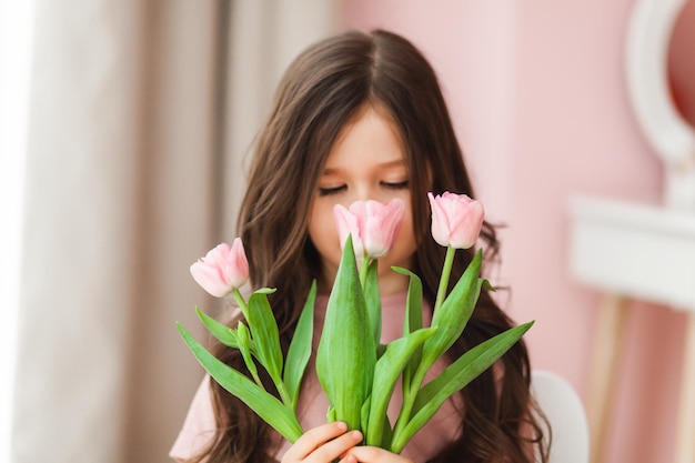 Portrait of a little girl with long dark hair closeup The baby hugs a bouquet of fresh delicate pink tulips A gift for the holiday spring time