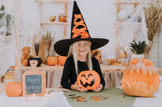 Photo portrait of a little girl with long hair in a witch costume with huge witch hat is holding a pumpkin