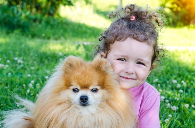 屋外で犬を抱いて幸せな少女の肖像画。美しい赤ちゃんとポメラニアンスピッツ。子供とペットが探しています