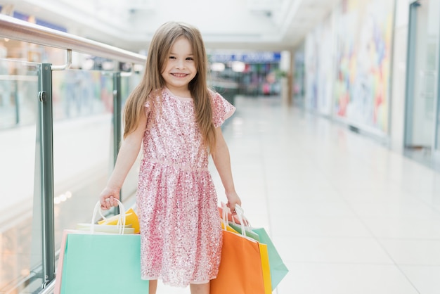 Ritratto di una bambina felice nel centro commerciale. una ragazza sorridente che ride in un abito rosa con borse colorate in mano è impegnata nello shopping. modello per la tua pubblicità