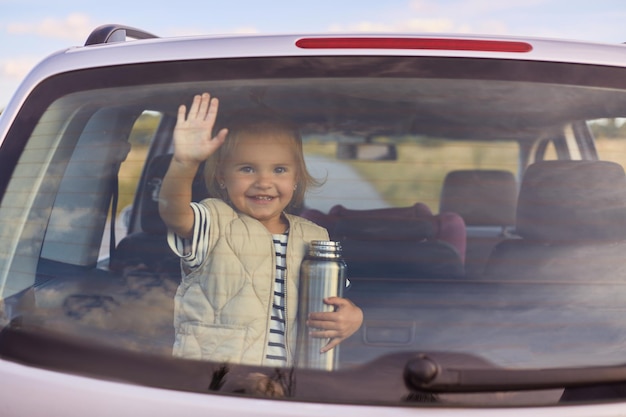 Portrait of little infant baby girl waving her hand into car window greeting or saying goodbye holding thermos in hands traveling by car with her family