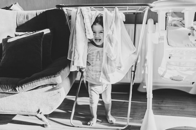 Portrait of a little pretty cute toddler girl adorable baby hangs laundry on clothesline and play on floor at home Black and white photo