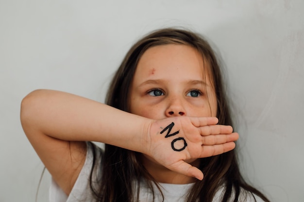 Portrait of little scared bruised girl raising hand to cover mouth showing inscription no on open palm