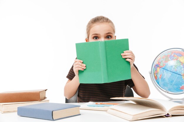 Portrait of little school girl reading studying books and doing homework while sitting at desk in class isolated over white wall