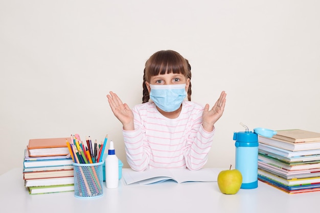 Ritratto di piccola studentessa con trecce che indossa una maschera protettiva seduta alla scrivania circondata da libri allarga le braccia in visita a scuola durante l'epidemia di virus influenzale