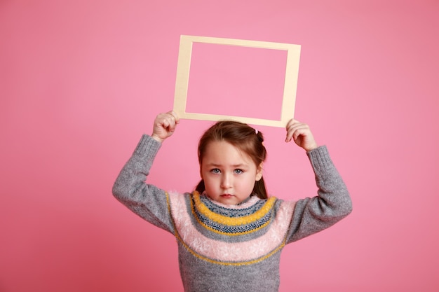 Portrait of a little smiling girl holding blank frame for mock-up on a pink background.