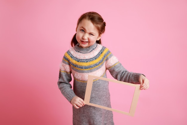 Portrait of a little smiling girl holding blank frame for mock-up on a pink background.