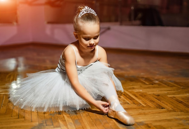 A portrait of a little sweet ballerina in a white tutu sitting on the floor and tying pointe shoe in the dance studio Children and ballet