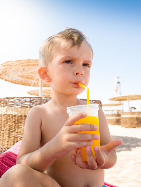 Ritratto di un bambino seduto sul lettino del sole sulla spiaggia del mare e che beve succo d'arancia dalla paglia