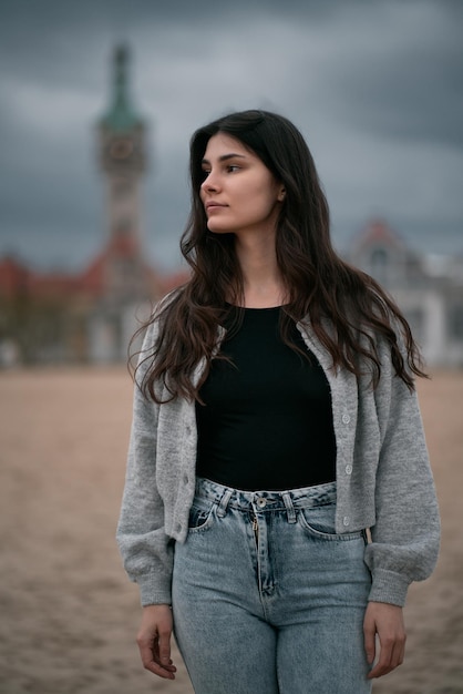 Portrait of long brown haired girl standing alone on the sand beach during cloudy weather Spring or autumn vacation on the seaside concept Close up portrait of caucasian woman