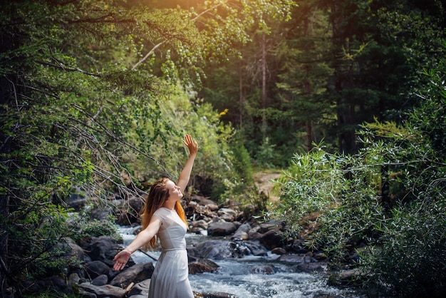Портрет длинноволосой брюнетки в белом платье позирует на фоне небольшой горной реки и зеленых деревьев. Красивая молодая женщина на берегу лесного ручья.