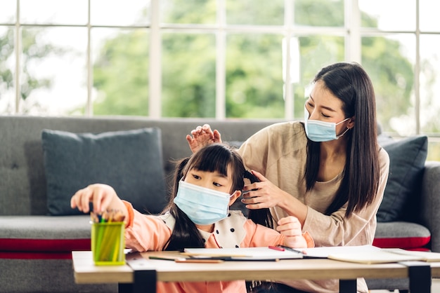 Portrait love asian family mother and little asian girl learning and writing in book with pencil making homework in quarantine for coronavirus wearing protective mask with social distancing at home