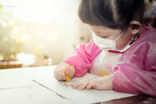 Portrait of lovely  Asian girl wearing facial hygienic mask  and drawing her art work with crayons ,selective focus. Quarantine,
Home isolation during COVID-19 pandemic.