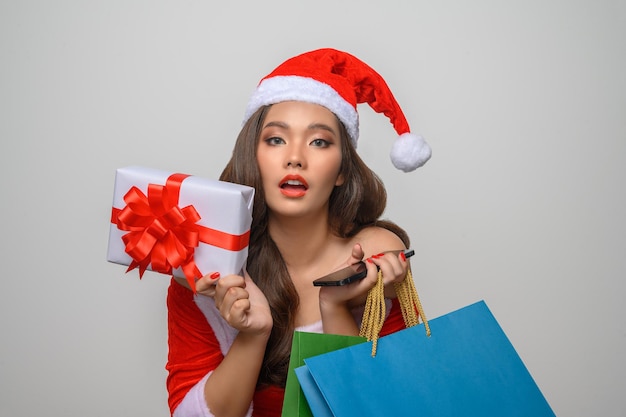 Ritratto di una bella donna asiatica in costume rosso di babbo natale in posa con borse della spesa, smartphone e confezione regalo in mano