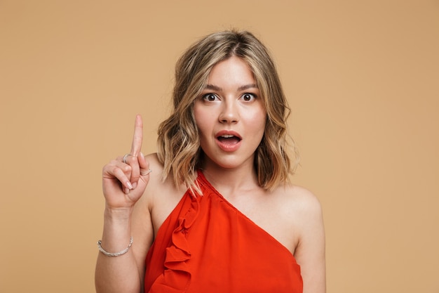 Portrait of a lovely blonde girl wearing dress standing isolated over beige wall, pointing finger